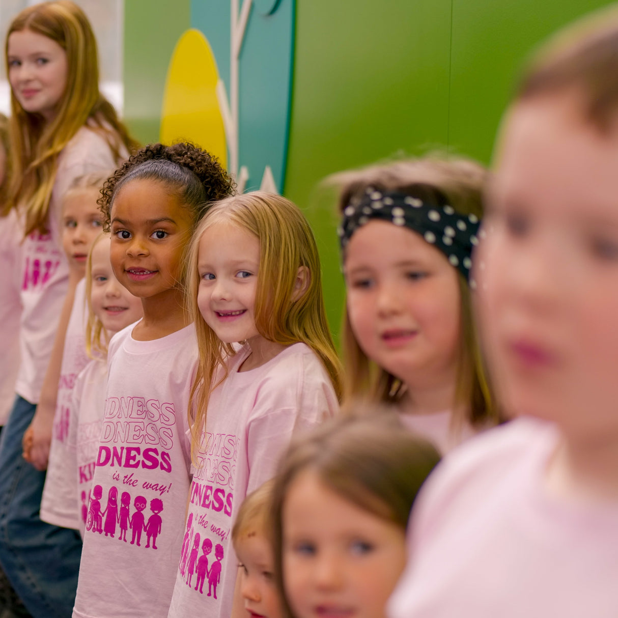 Children wearing pink kindness t-shirts, standing in a line against a green wall.